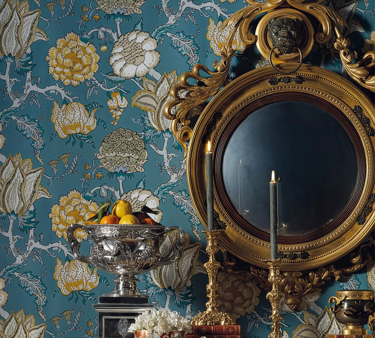 Lit taper candles in gold candlesticks before a gilded round mirror on teal floral wallpaper